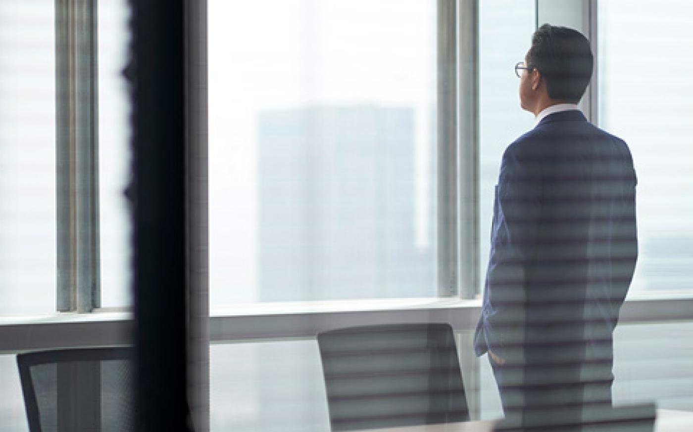 Business professional in a suit standing by office window, looking at city skyline in a modern workspace.